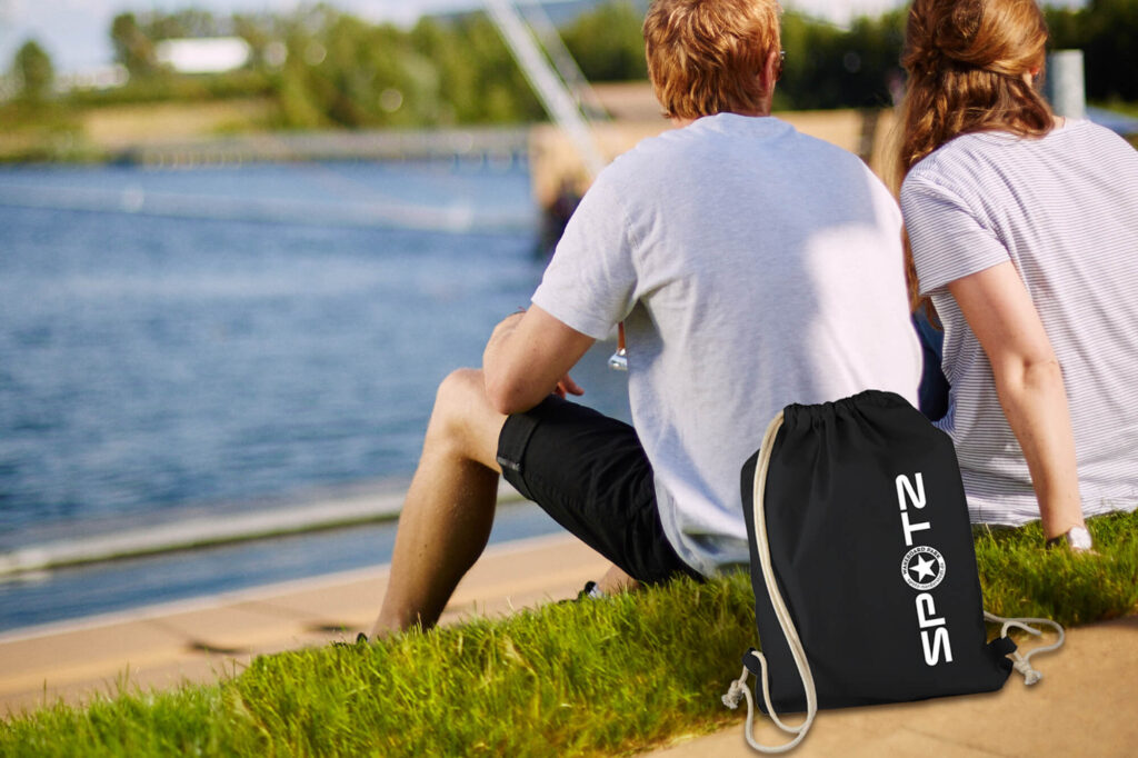 Sportbeutel mit SPOTZ Logo liegt auf dem Rasen hinter dem Rücken eines jungen Erwachsenen, der im Spotz Stadtpark Norderstedt auf dem Rasen vor der Wakeboardanlage sitzt